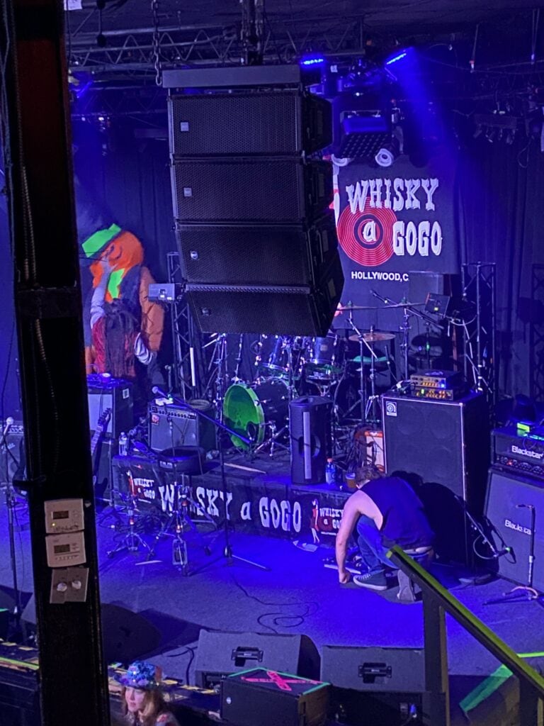 A stage at Whisky a Go Go in Hollywood, set up for a live performance. A person in a large leprechaun costume stands beside the drum set, while another person kneels, adjusting equipment under purple lighting.