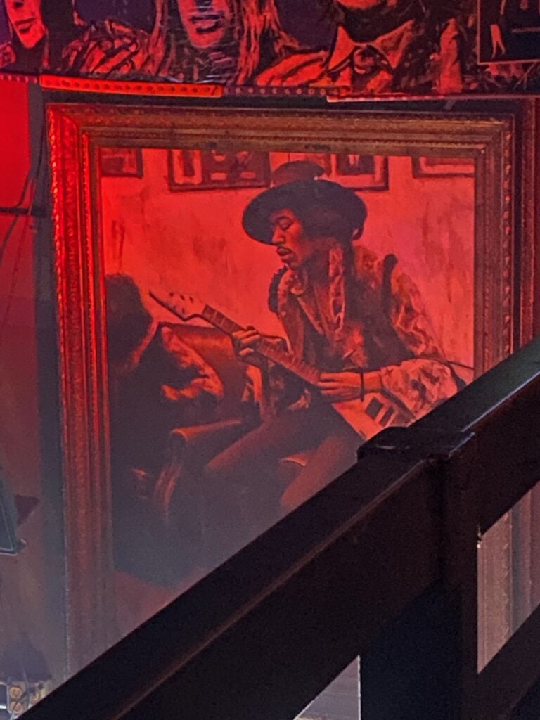 A large, framed painting of a man in a hat playing an electric guitar hangs on a red-lit wall, partially obscured by a railing, with mural-style portraits above it.