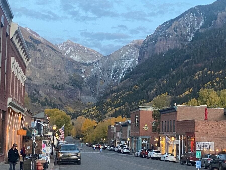 A small mountain town street at dusk, lined with brick buildings and shops, with cars parked along the road. Snow-capped mountains and autumn trees rise in the background—perfect for exploring the many things to do in Idaho Springs, CO.