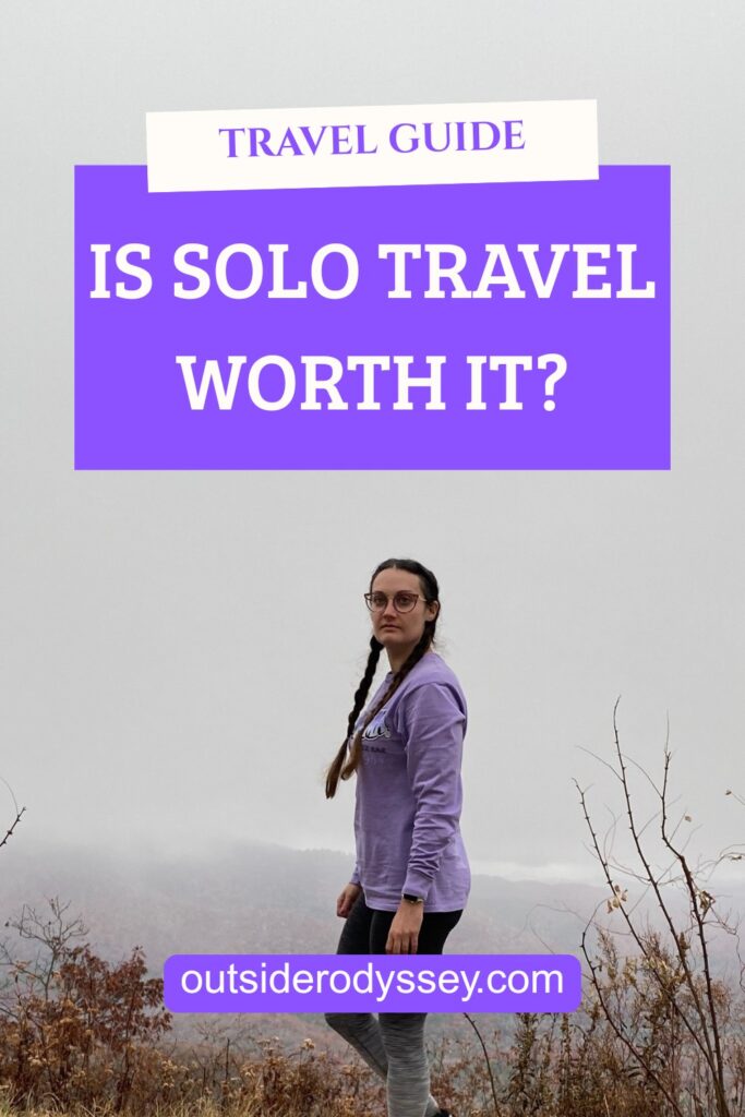 A woman in a purple sweatshirt stands on a hill with foggy scenery in the background. Text reads: Is Solo Travel Worth It? Find your answer at outsiderodyssey.com with our travel guide.