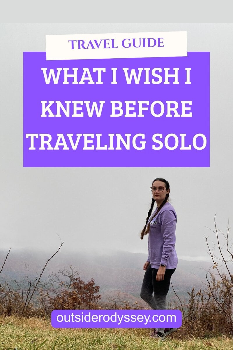 A woman in a purple outfit stands outdoors on a cloudy day with foggy hills behind her. Text on the image reads: “What I Wish I Knew Before I Took My First Solo Trip. outsiderodyssey.com. Travel guide.”.