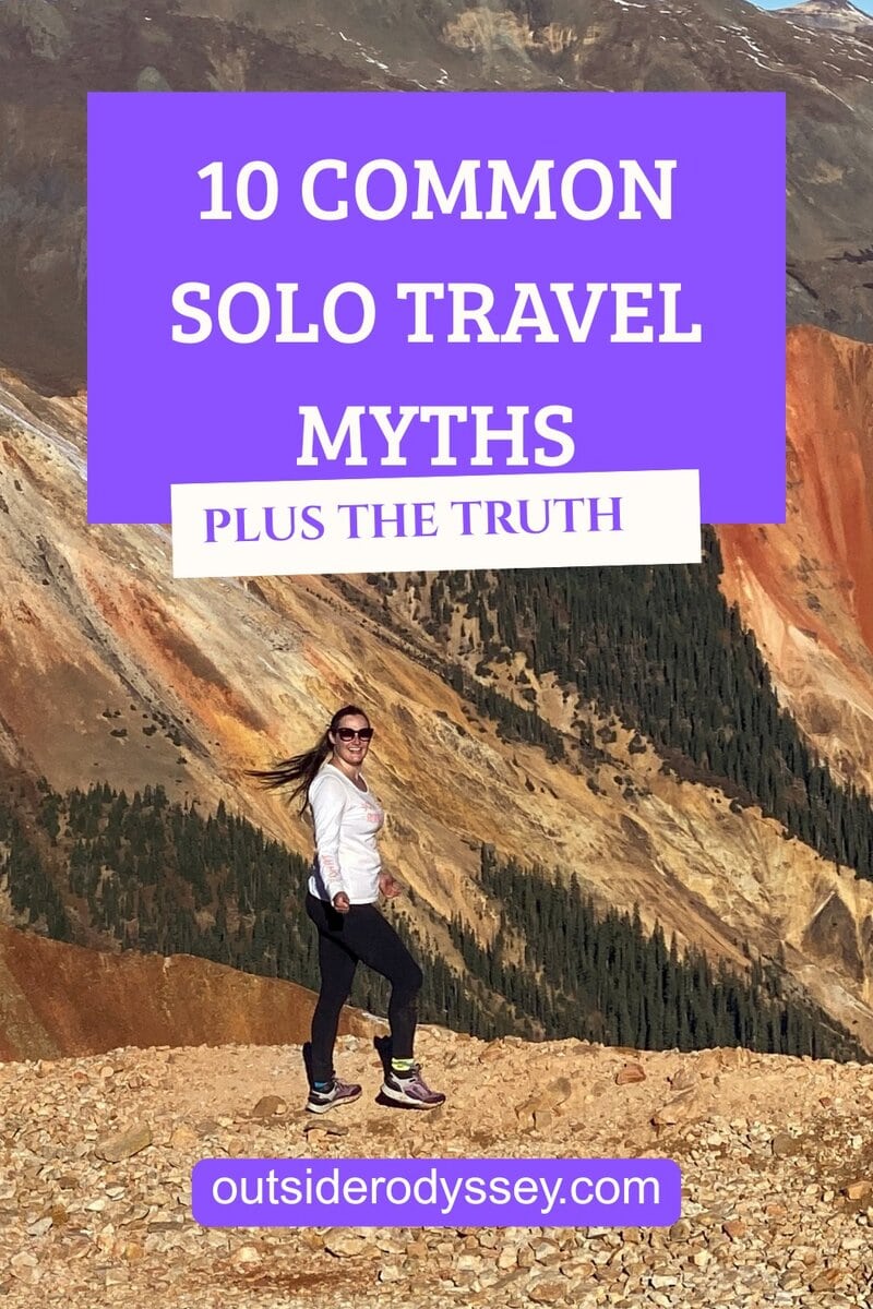 A woman stands on a rocky mountain trail with colorful slopes and trees behind her. Text reads: 10 Common Solo Travel Myths Debunked and outsiderodyssey.com.