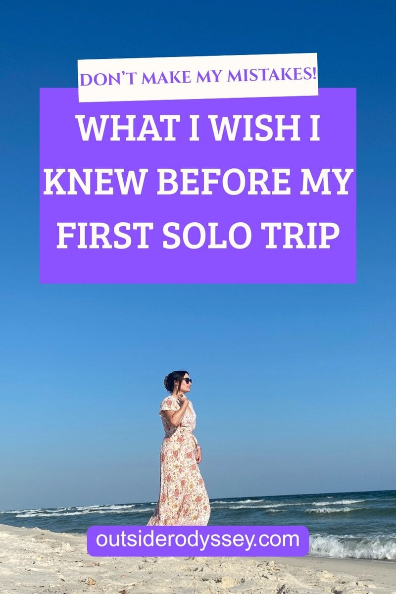 A woman in a floral dress stands on a sandy beach beneath a clear blue sky; overlaid text reads, DONT MAKE MY MISTAKES! WHAT I WISH I KNEW BEFORE MY FIRST SOLO TRIP and outsiderodyssey.com.