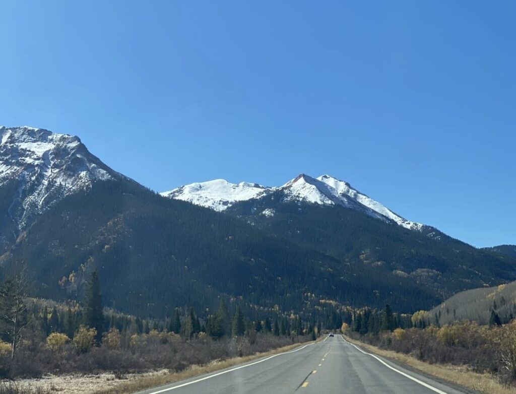 A paved road leads toward distant snow-capped mountains under a clear blue sky, with forests and patches of autumn foliage on either side.