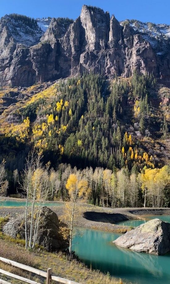 A turquoise river flows past rocks and autumn trees with yellow leaves, set below a forested slope and tall rocky mountains dusted with snow under a clear sky. A wooden fence runs along the foreground.