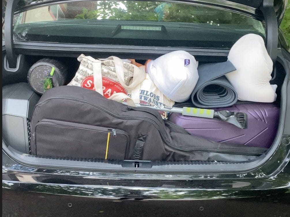 The open trunk of a car is packed with various items, including bags, a neck pillow, a yoga mat, a foam roller, a suitcase, and a rolled-up newspaper.