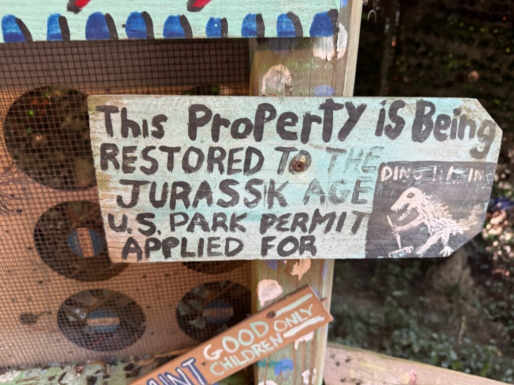 A hand-painted wooden sign at Abita Mystery House reads, This property is being restored to the Jurassic Age. U.S. park permit applied for, with a dinosaur drawing and DINO LAND label, surrounded by colorful painted decorations.