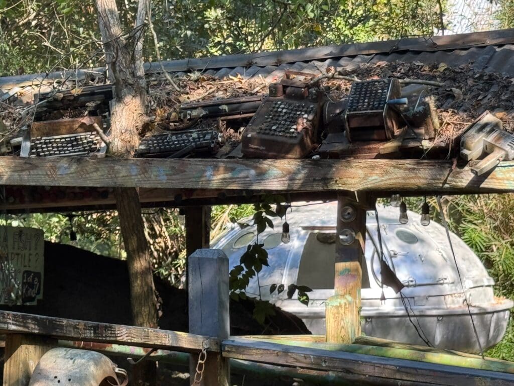 Several old typewriters covered in leaves rest on a weathered wooden roof in a wooded area, with the Abita Mystery House’s metallic dome-shaped structure visible among the trees in the background.