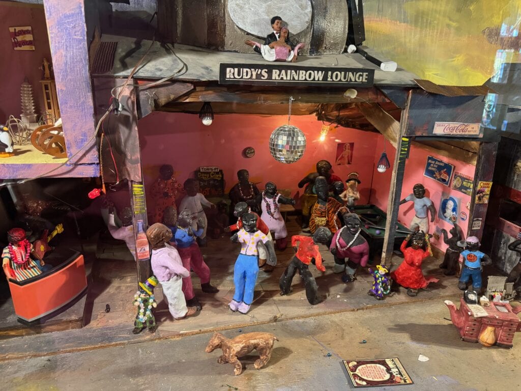 A detailed miniature diorama of Rudy’s Rainbow Lounge shows figures dancing, playing music, and socializing inside a lively, colorful bar. A disco ball hangs from the ceiling and a dog stands outside the entrance.