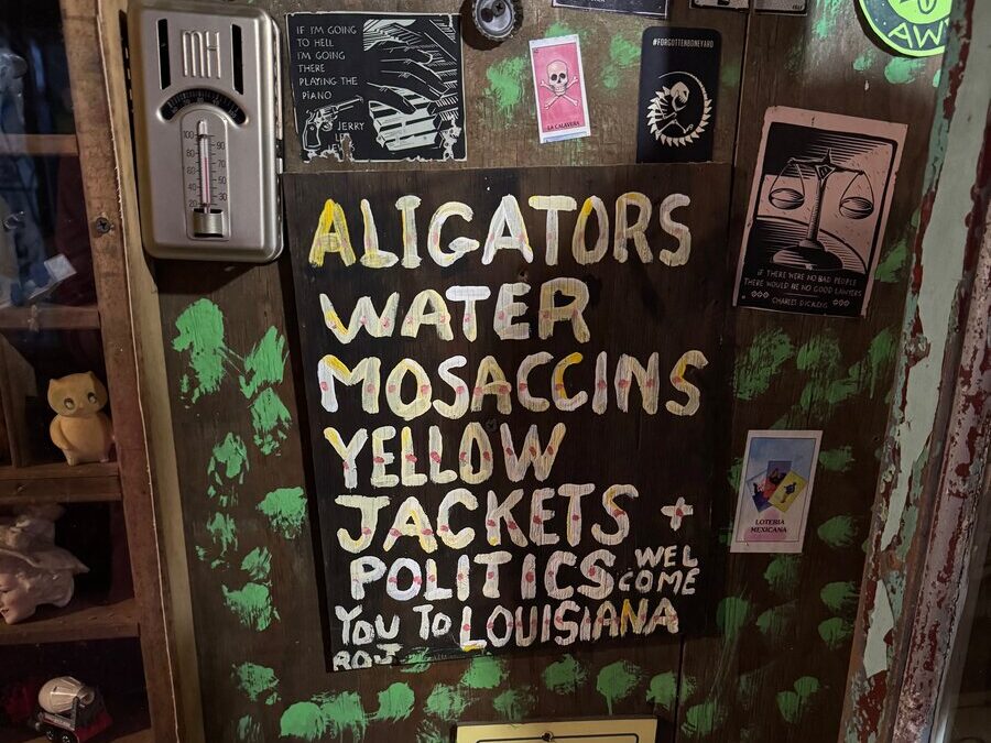 A wooden wall at the Abita Mystery House features hand-painted text listing “Alligators, Water Moccasins, Yellow Jackets + Politics” to warn visitors. Surrounding it are eclectic stickers, posters, and objects welcoming you to Louisiana.
