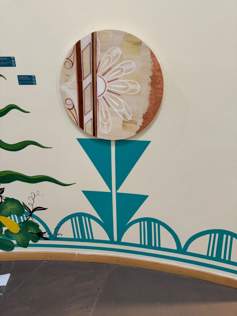 A circular artwork with abstract patterns is mounted on a cream wall featuring teal geometric designs and green leafy accents near the bottom left. The floor is partially visible.