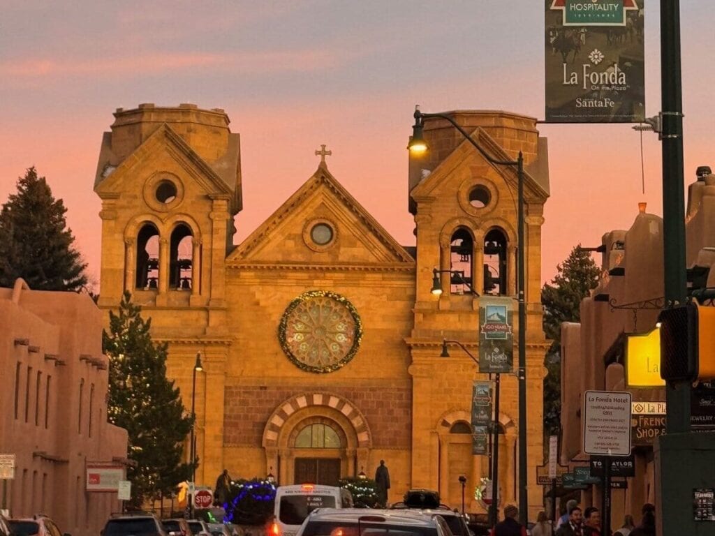A street scene at sunset in Santa Fe, New Mexico, features the illuminated facade of a church with twin towers and a large wreath. Cars and people line the street, and a banner for La Fonda Hotel hangs on a lamppost.