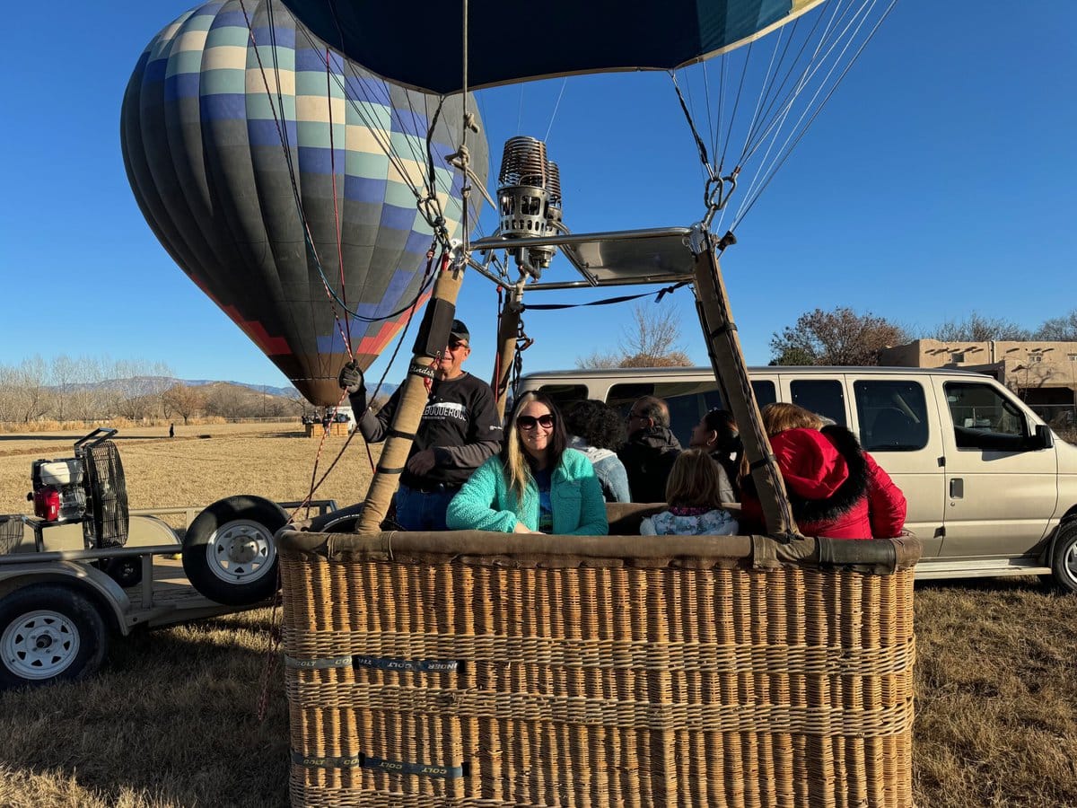 A group of people sits in a hot air balloon basket on the ground, next to a white van and trailer. Another inflated hot air balloon is visible in the background under a clear blue sky.