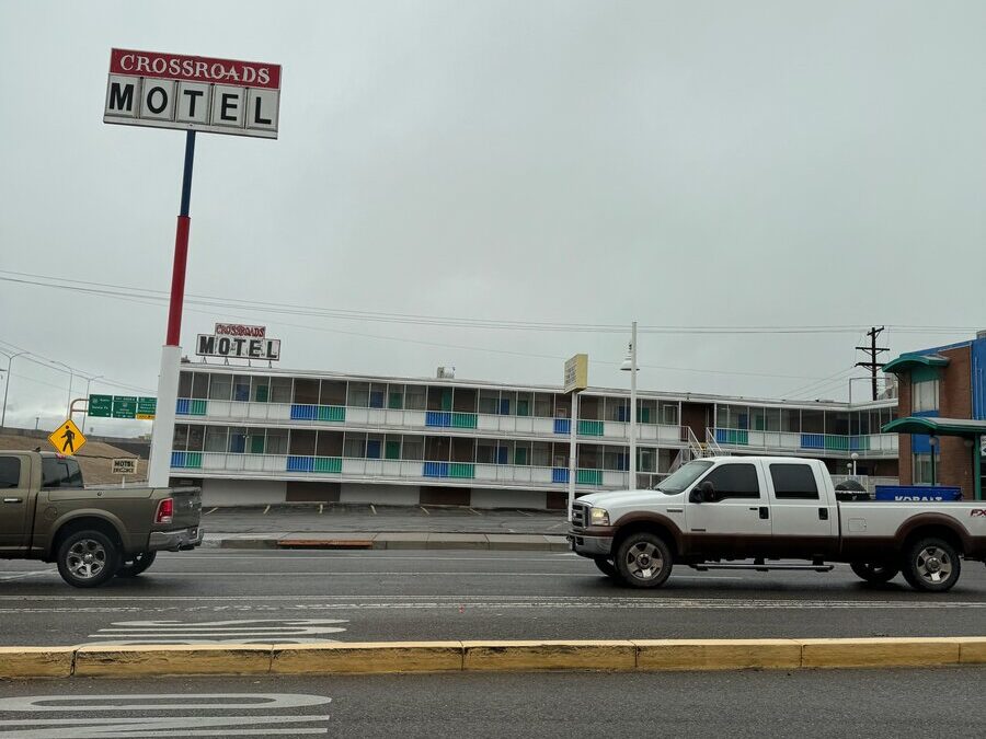 A street view of the Crossroads Motel with two pickup trucks driving by on a cloudy day. The motel is a three-story building with colorful balcony railings and a large sign in front.