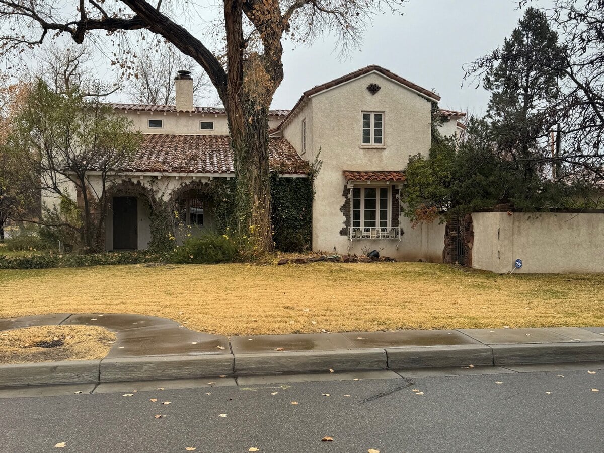 A two-story beige stucco house with a red-tile roof sits behind a patchy yellow lawn and sidewalk. Large leafless trees stand in front, and the sky appears overcast.