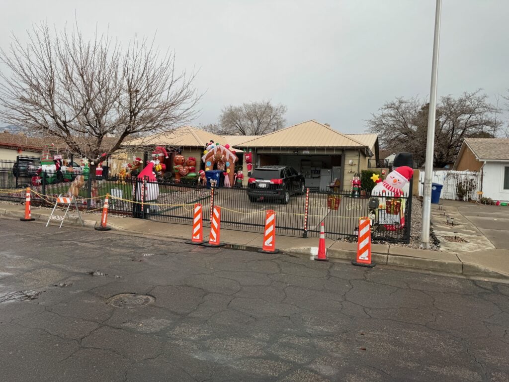 A house decorated with large inflatable Christmas figures, including Santa and a gingerbread house, is surrounded by orange traffic cones and caution tape. A black truck is parked in the driveway. Trees are bare of leaves.