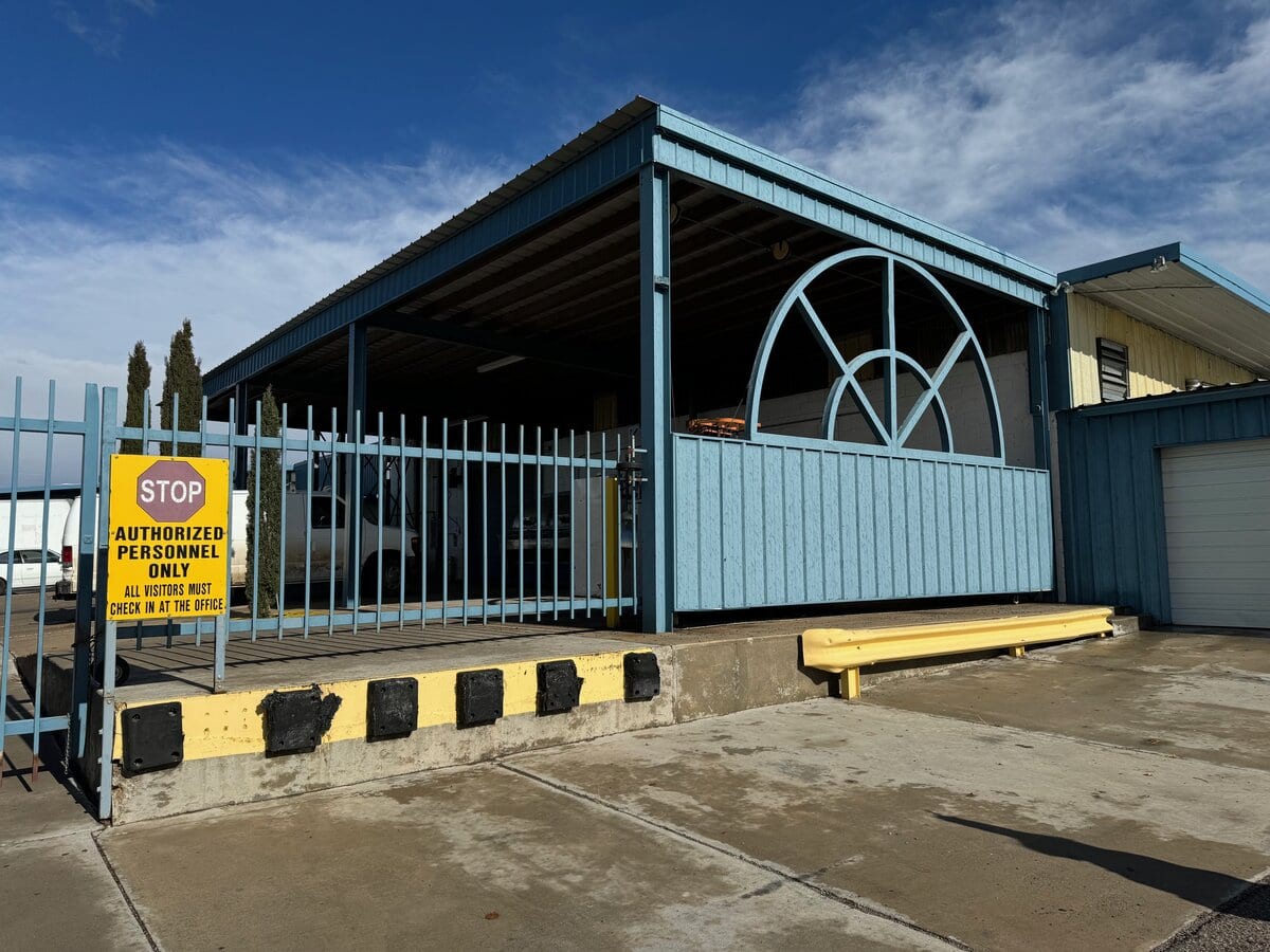 A blue industrial building with a large metal wheel structure on its side is behind a blue fence. A yellow sign on the fence reads STOP AUTHORIZED PERSONNEL ONLY. The sky is partly cloudy.
