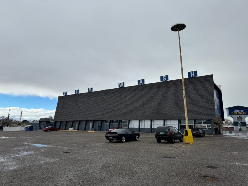 A large, dark-roofed building with CAR WASH letters on top sits in a mostly empty parking lot under a cloudy sky. Several cars are parked, and a tall, weathered lamppost stands in the foreground.