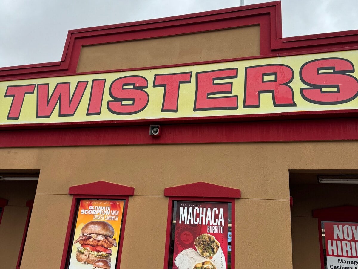 A restaurant with a large sign reading TWISTERS above posters advertising a Machaca Burrito and other menu items. A Now Hiring sign is partially visible on the right. The building is tan with red trim.