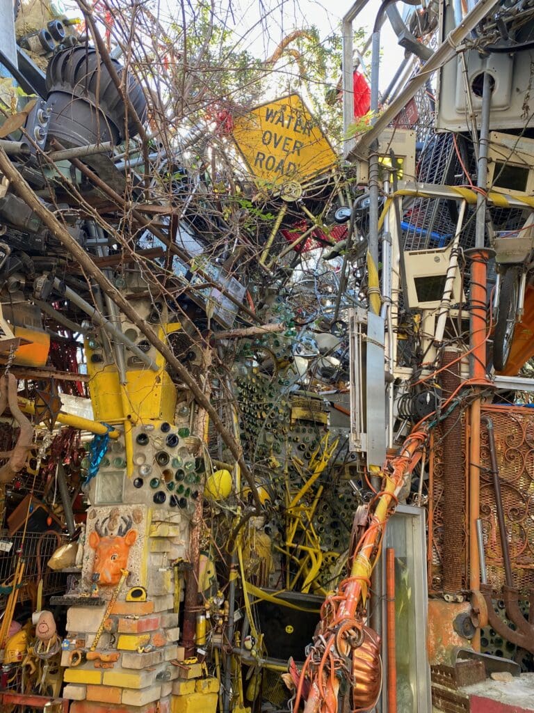 A dense, colorful jumble of pipes, wires, metal, and found objects forms an intricate outdoor sculpture. A yellow “Water Over Road” sign is visible among branches and industrial materials.