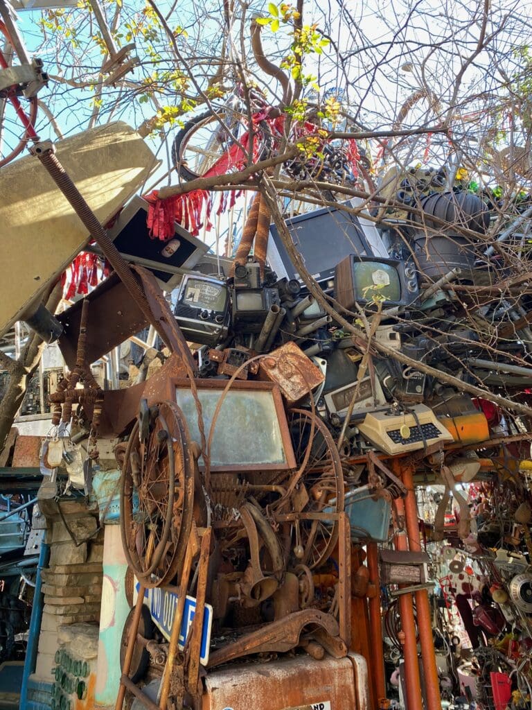 A dense jumble of old electronics, TVs, typewriters, and metal parts stacked outdoors among tree branches, with sunlight filtering through and red ribbons hanging from the branches.