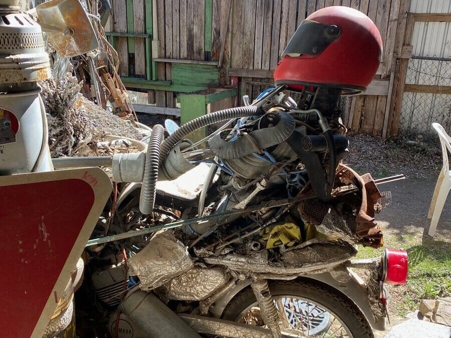 A motorcycle sculpture with a mannequin wearing a red helmet, made from various metal parts and pipes, is displayed in a yard with wooden fencing and assorted outdoor items in the background.