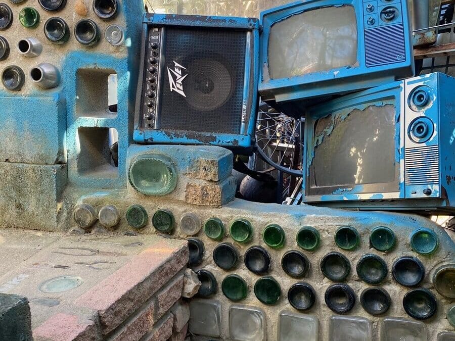 A wall made of glass bottles and bricks is decorated with old blue TVs and a speaker. Sunlight filters through the scene, and part of an American flag is visible at the top left.