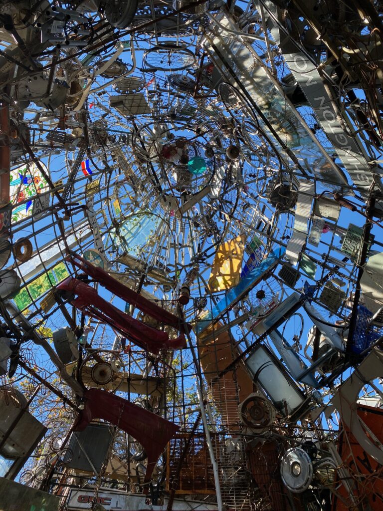 Looking up through a dome made of eclectic, recycled materials like metal, glass, and colorful signs, with blue sky and sunlight visible through gaps, creating a kaleidoscopic, artistic ceiling.
