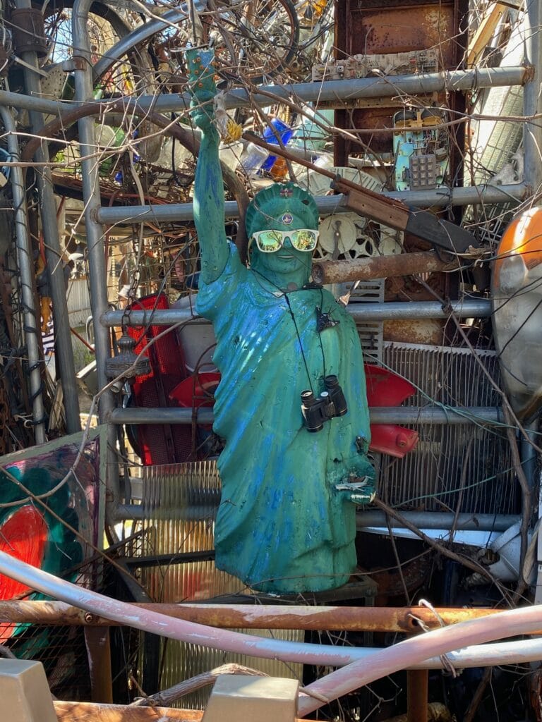 A small Statue of Liberty replica wearing sunglasses and binoculars stands among a chaotic arrangement of metal, wires, and assorted objects in an outdoor art installation.