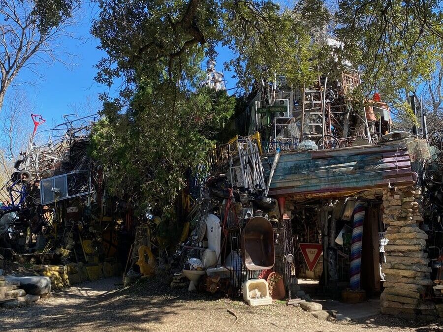 An eclectic outdoor art installation made of various objects, metal, wood, and found items, sits beneath large leafy trees in bright daylight, with a small rustic structure in front.
