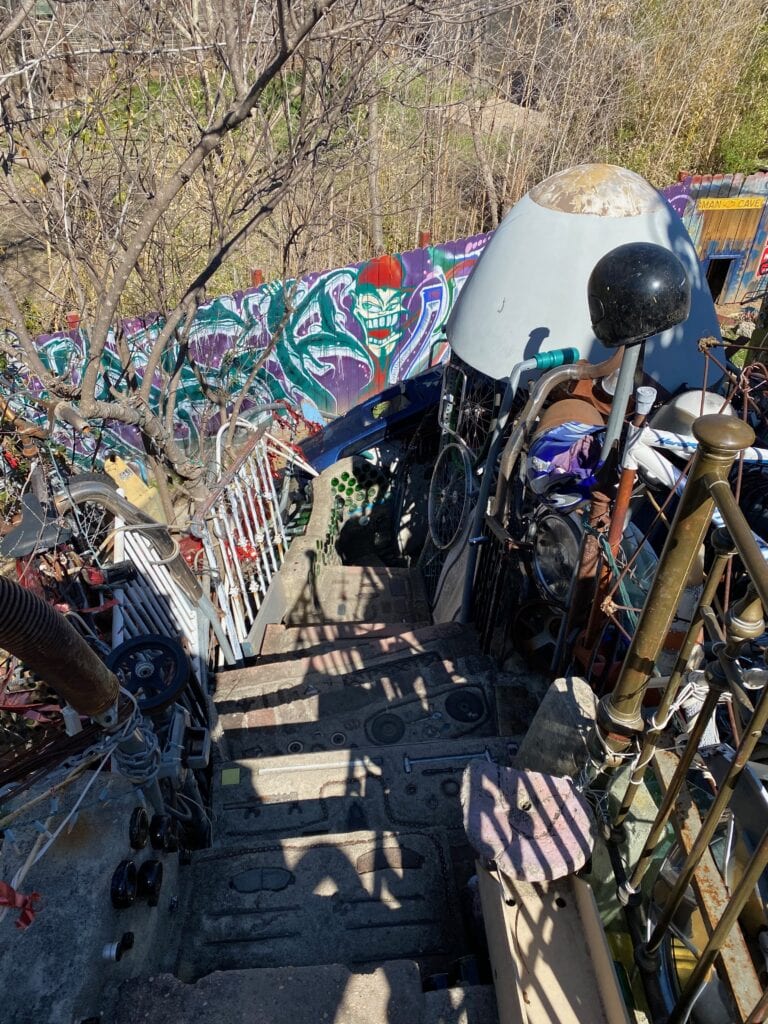 A narrow outdoor staircase lined with eclectic objects leads down past colorful graffiti on a wall. There are trees, a bicycle, and various metal and plastic items cluttering the area, creating an artistic, chaotic scene.