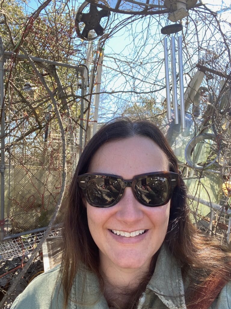 A woman wearing sunglasses smiles outdoors in front of metal structures and leafless branches under a sunny blue sky.