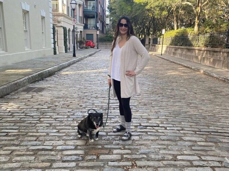A woman wearing sunglasses and casual clothes stands on a cobblestone street holding a leash attached to a small black and white dog sitting beside her. Trees and buildings line the street in the background.