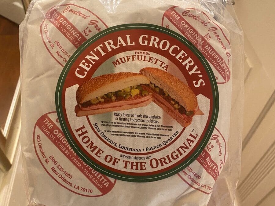 A hand holds a wrapped package from Central Grocery’s, featuring a label for their famous muffuletta sandwich—one of the must-try cheap eats in New Orleans. The round label shows a sandwich illustration and reads Home of the Original with store details.