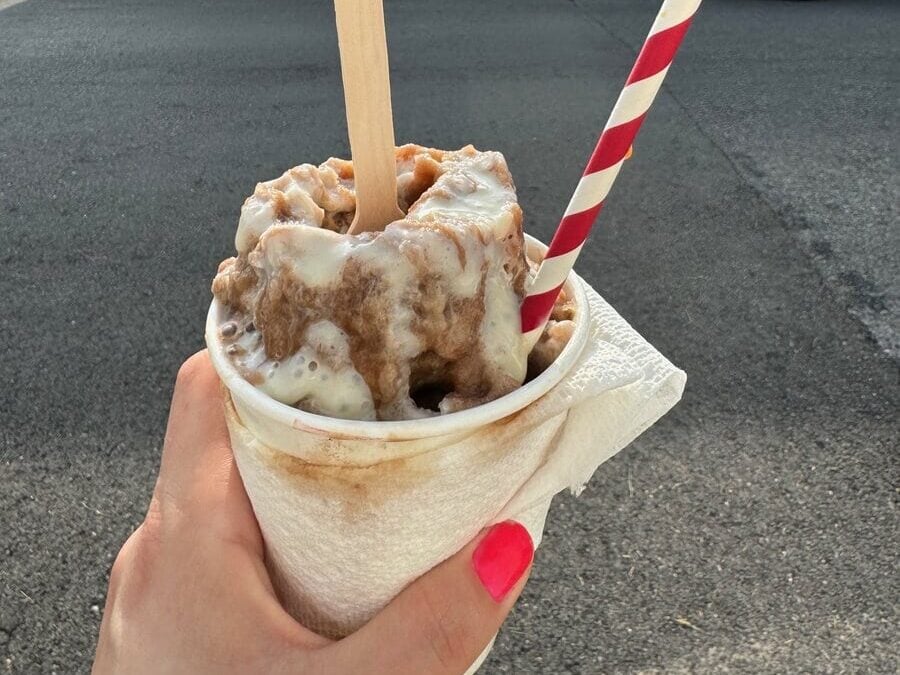 A hand with bright pink nail polish holds a cup of melting ice cream—a classic find among cheap eats in New Orleans—with a wooden spoon and red-striped straw, set against a colorful mural of two people in elaborate headdresses.