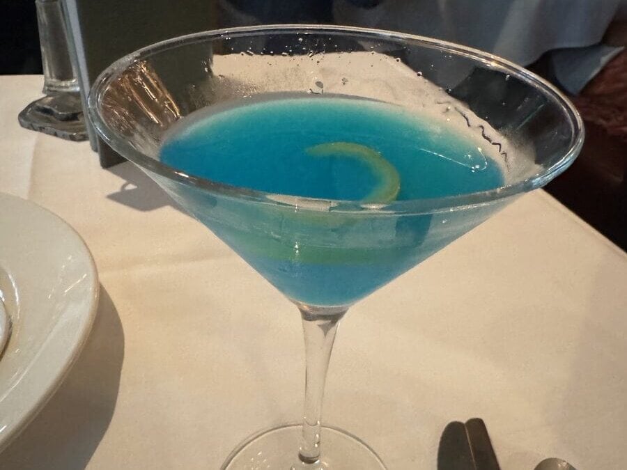 A martini glass filled with a vibrant blue cocktail, garnished with a twist of lime, sits on a white tablecloth next to a fork and spoon in a lively restaurant known for cheap eats in New Orleans.