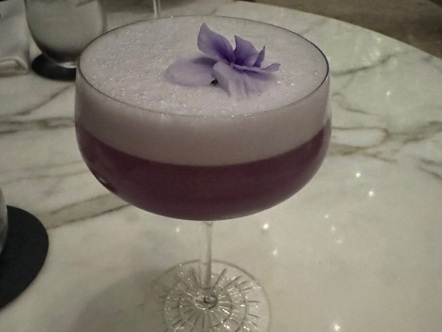 A frothy purple cocktail in a coupe glass, garnished with a light purple flower, sits on a marble table—perfect for sipping after exploring cheap eats in New Orleans.