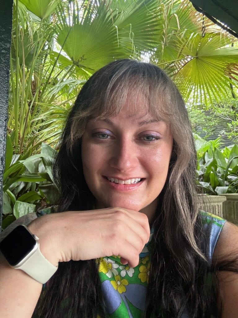 A woman with long brown hair and bangs smiles at the camera. She is wearing a colorful top with a floral pattern and a white smartwatch—perfectly dressed for exploring cheap eats in New Orleans. Lush green tropical plants fill the background.
