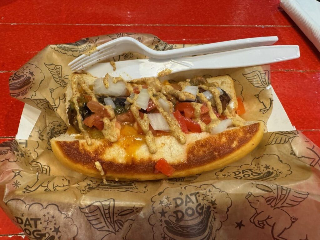 A hot dog topped with diced tomatoes, onions, and other vegetables sits on branded brown paper with a white plastic fork and knife, on a shiny red table.