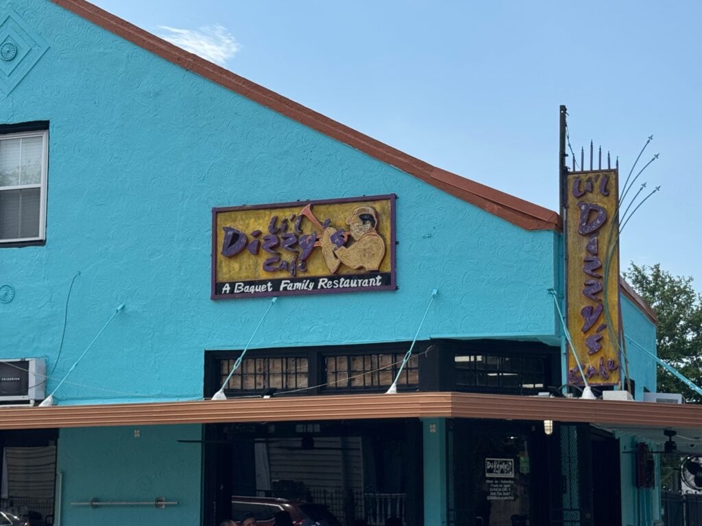 A turquoise building with a sign reading Lil Dizzy’s Cafe: A Baquet Family Restaurant in yellow and purple, known for cheap eats in New Orleans. The sign features an illustration of a man in a hat, with a matching vertical sign at the corner.
