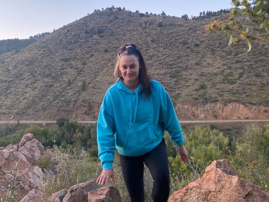A woman in a bright blue hoodie and black leggings climbs over rocks outdoors, with a grassy hillside and clear sky in the background. She is smiling and wearing sunglasses on her head.