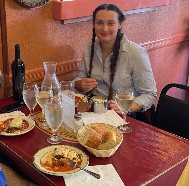 A woman with braided hair sits at a restaurant table set with food, wine, and glasses of water. Behind her is a decorative mirror featuring an image of a woman holding scales. The setting is cozy and warmly lit.
