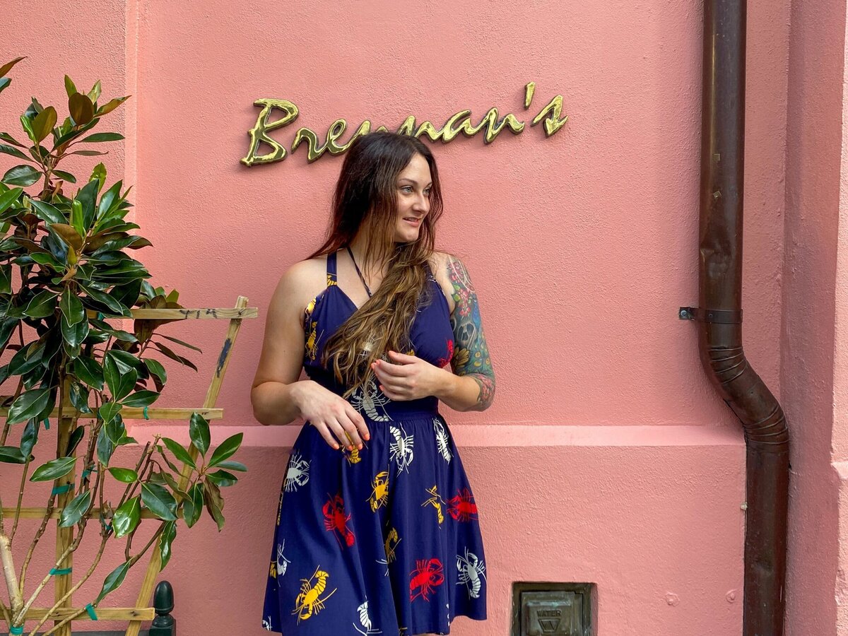 A woman in a blue dress with colorful prints stands smiling against a pink wall with gold cursive text reading Brennans. A plant is on her left and a dark drainpipe runs down the right side of the wall.