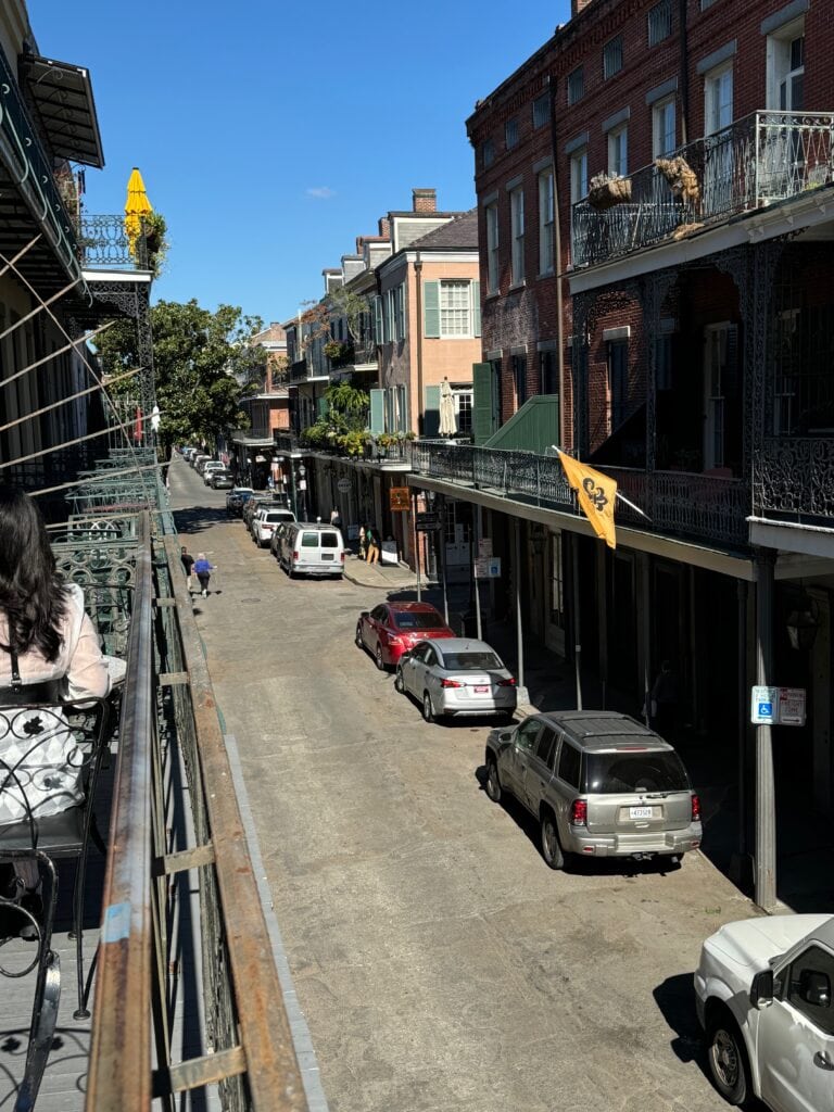 A sunny street view in the French Quarter of New Orleans shows parked cars, historic buildings with wrought-iron balconies, and a yellow flag waving above the sidewalk. People are sitting on a balcony overlooking the street.
