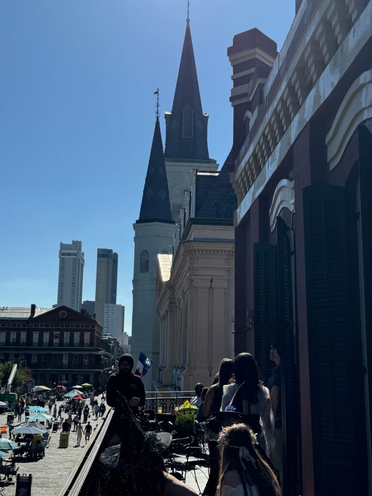 People stand on a balcony overlooking a lively street scene with shops, market stalls, and a park. In the background, there is a tall church with spires and modern buildings under a clear blue sky.