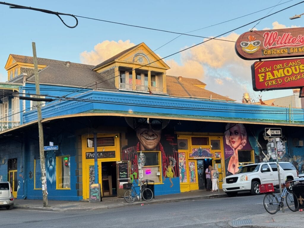 A vibrant corner building painted blue and yellow houses “Willie’s Chicken Shack,” a go-to spot for cheap eats in New Orleans. Murals of faces decorate the walls, while people stand near the entrance on a sunny day.