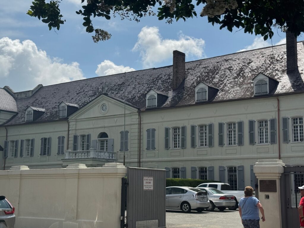 A large, white, two-story building with gray shutters and a dark roof, seen on a sunny day. Several cars are parked in front, and two people walk along the sidewalk in the lower right corner.