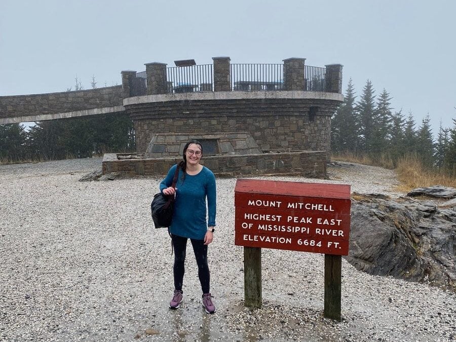 Woman drenched in rain water posing by Mt. Mitchell sign at the summit of the trail.