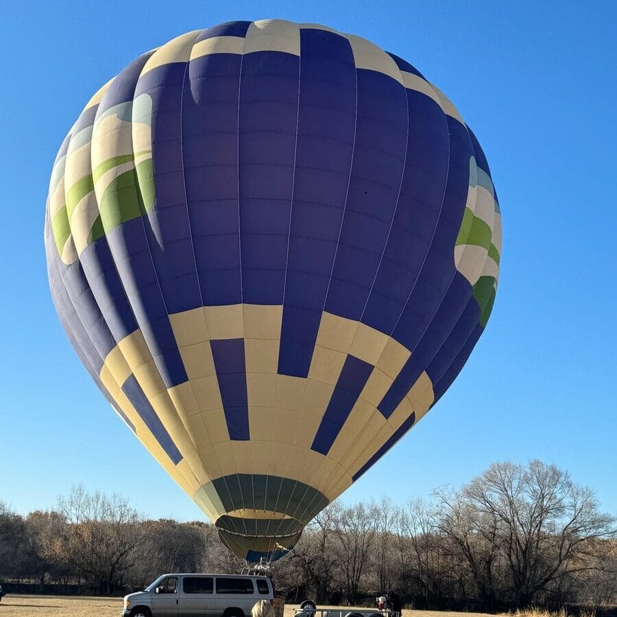 A large purple, cream, and green hot air balloon is being prepared for riding in a hot air balloon in a grassy field. A person stands nearby, and a white van with a trailer is parked under the clear blue sky.