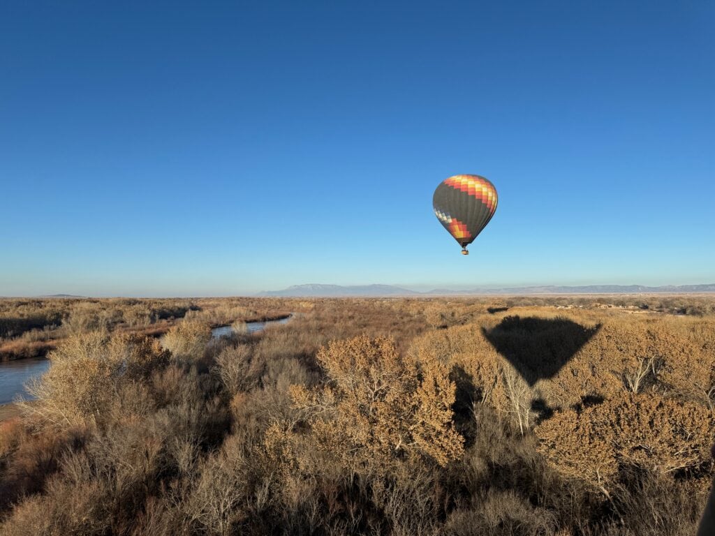 A colorful hot air balloon floats above a forest of autumn trees, casting a large shadow on the foliage below as if you were riding in a hot air balloon. A river winds through the peaceful landscape under a clear blue sky.